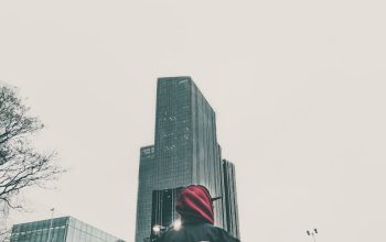 Individual in streetwear looks at city skyscraper, showcasing urban fashion against a modern cityscape.