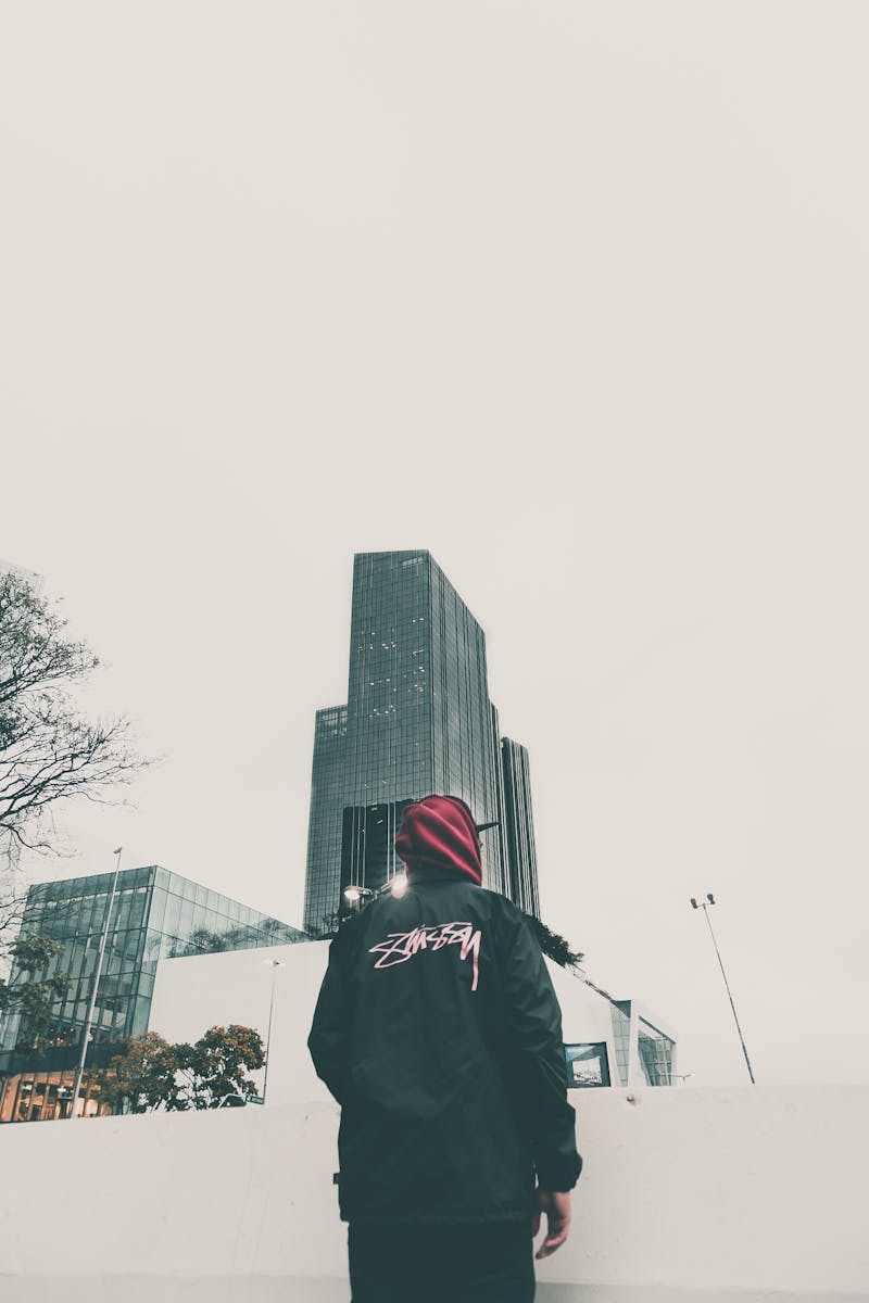 Individual in streetwear looks at city skyscraper, showcasing urban fashion against a modern cityscape.